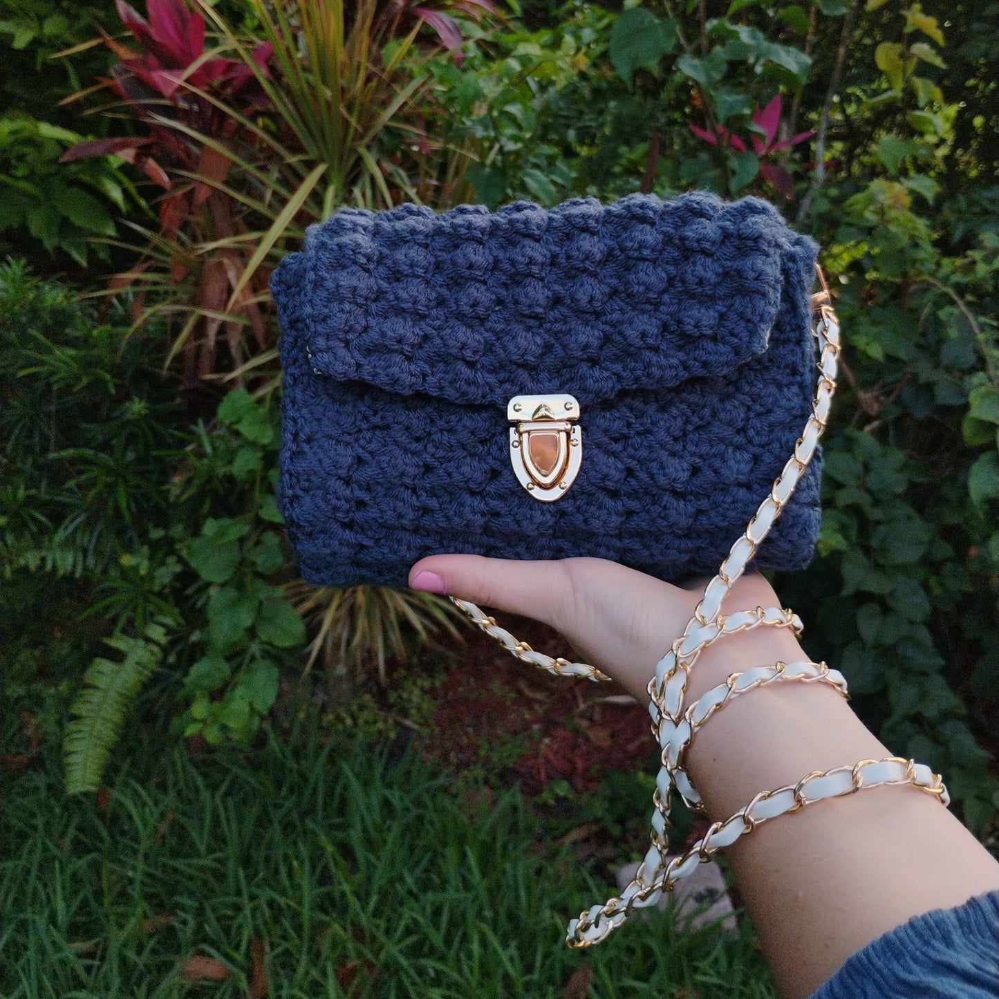 Navy blue crochet bag held by a hand with a gold clasp against a green foliage background
