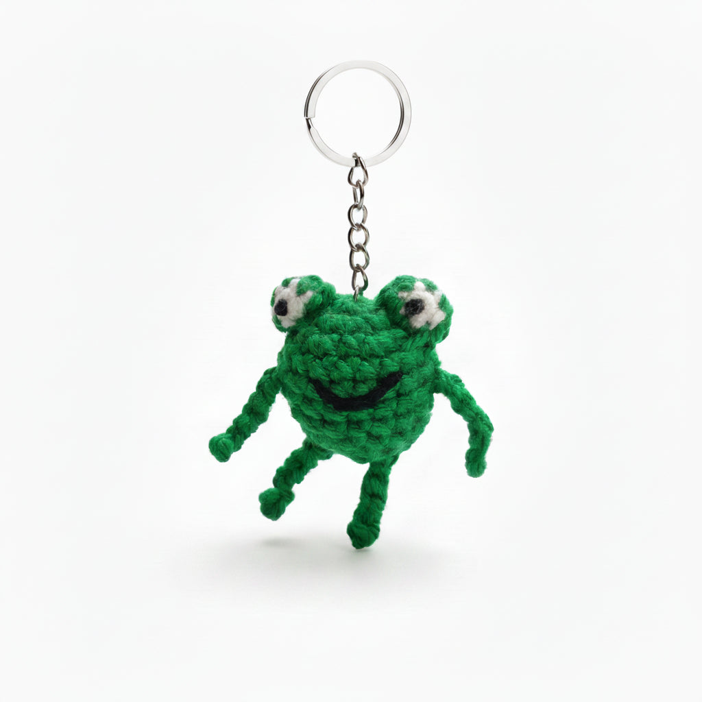 Green crocheted frog keychain hanging on a chain against a blurred natural background.