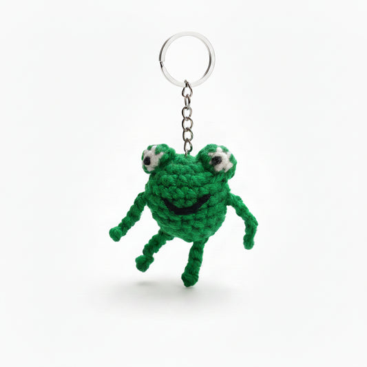 Green crocheted frog keychain hanging on a chain against a blurred natural background.