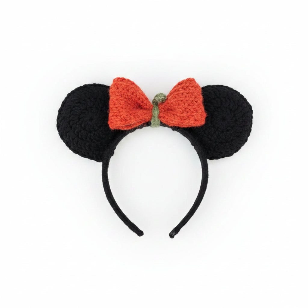 Hand holding a crocheted black mouse ears headband with a pumpkin bow on a pebbled surface.