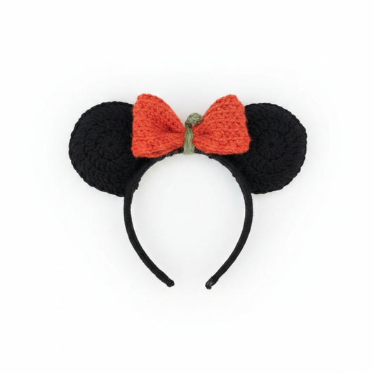 Hand holding a crocheted black mouse ears headband with a pumpkin bow on a pebbled surface.