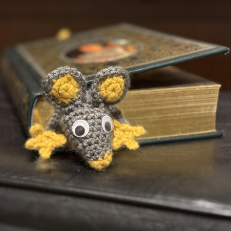 Crocheted mouse bookmark with yellow feet and gray body on a dark surface in a book